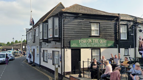 People are sat outside a building which is branded with a green Fish on the Quay sign. It is a sunny day and the road beside the quay is on the left hand side.
