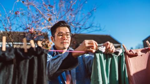 A man hangs out laundry on the line, the sky behind him is bright blue.
