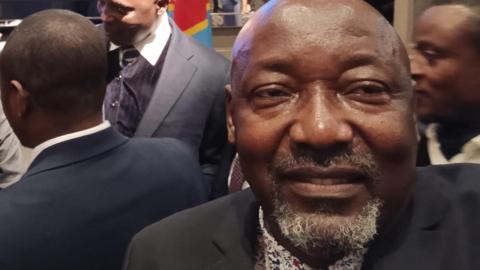 Jacques takes a selfie in a crowded room. He is smiling and is wearing a suit. He is bald and has a silver stuble.