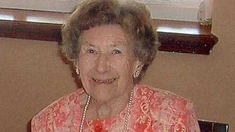 Una Crown with short curly light brown hair and wearing gold earrings and a pearl necklace and a coral and light pink patterned top. She is smiling and sitting down.