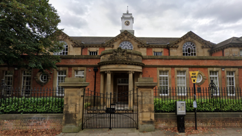 The exterior of Newcastle's Royal Grammar School which is a grand, red-bricked building with stone columns, large arched windows and a clock tower. It is behind a black metal fence with gold lettering on gate reading: "Newcastle Royal Grammar School". There is a payment machine for parking out front.
