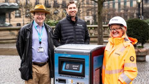 Two men and a woman stand beside a smart waste bin in Sheffield city centre. The man on the left has dark brown hair and glasses, and wears a brown fedora hat, black leather jacket, purple shirt and beige chinos. The man in the middle has short brown hair and a beard, and wears a black puffer gilet and black long sleeved top. The woman has blonde hair and glasses, and wears a white hard hat and fluorescent orange jacket. 