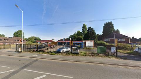 A set of railings with an industrial-looking compound behind where there are buildings with signage for a hand car wash and some parked cars.