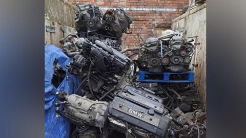 A pile of dirty car parts. They are in a compound with high walls on three sides, concrete at the sides and brick at the back.