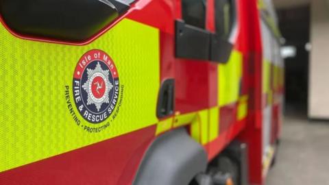 A closeup of the side of a fire engine, which is red and yellow in colour and features the Isle of Man Fire and Rescue Service logo.