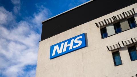 A grey building with an NHS logo on the side. We can see some windows. There is a blue sky behind it with some white clouds. 