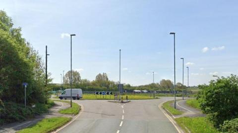 The approach to a roundabout. There is a white van exiting the roundabout. There is pavement on both sides and a crossing as the dropped kerb road approaches the junction.