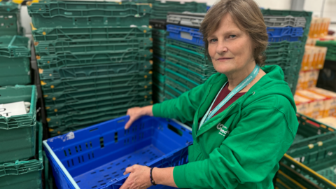 The photo shows a lady with a neutral expression looking at the camera. She is wearing a green hoodie, and is holding an empty blue crate. Behind her are stacks of crates about 5ft (1.5m) high which are also all empty.