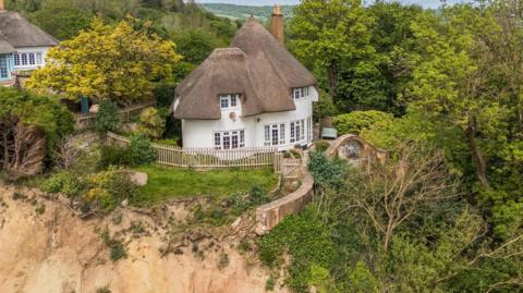 A picture looking from above down at the property and the cliff top. It is a large White House, with a brown roof. There is grassland and trees around the property.