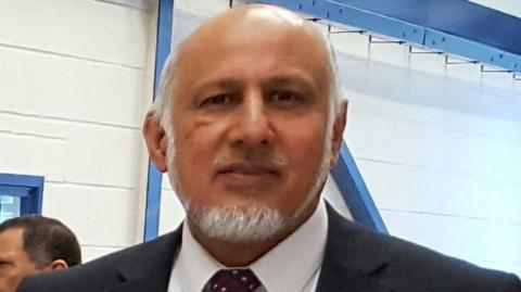 A man of Pakistani heritage with grey facial hair pictured from the shoulders up, wearing a dark coloured suit, white shirt and tie.