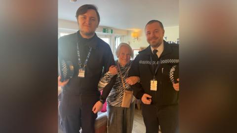 An elderly woman with a black and white top with a bag around her shoulder standing between two police officers in uniform. She is smiling and has her arms round their arms. The two male officers are also smiling facing the camera. Both are much taller than she is.