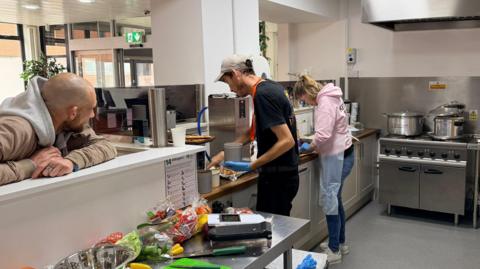 Two people working in a kitchen with a stove in the background and a man leaning on the counter