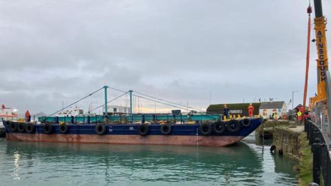 The bridge is lying on a barge in the harbour. The crane is on the quay. There are people wearing high-viz jackets and trousers and helmets on the barge and quay. The sky is cloudy.