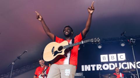 Myles Smith performing at Radio 1's Big Weekend in 2024. He has his arms in the air and is looking out towards the crowd with his guitar round his neck. There is an illuminated BBC Introducing sign behind him. 