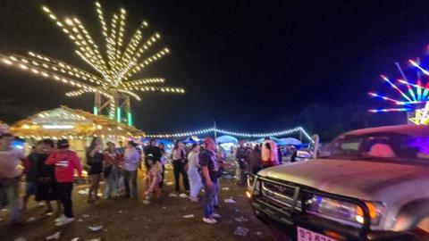 A crowd of people stand in the dark beside a car, debris on the grass and lights at a fair behind them