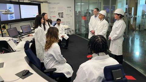 Sir Sadiq Khan is wearing a white hard hat and white lab coat, stood inbetween a man and a woman, both wearing lab coats and the woman also wearing a white hard hat. They are stood in front a group of six people all wearing white lab coats. Behind them is a set of screens. 