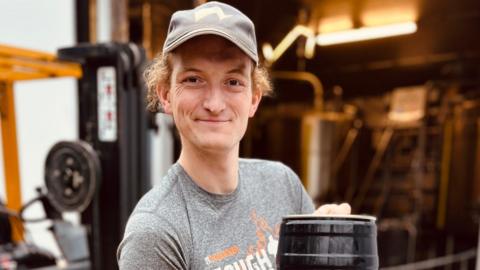 Harry Baynhams wearing a grey t shirt and cap. He has curly blonde hair under the cap. He is holding a black keg. Out of focus behind him is a brewery.