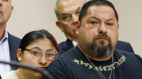 A woman leans against a man's shoulder as they listen to a press conference