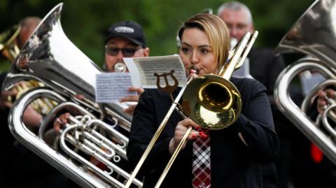 A woman playing a brass instrument. She is a member of a marching brass band which is playing in a parade during the traditional Whit Friday band contest in Manchester in 2024. Behind her can be seen two men who are also playing brass instruments