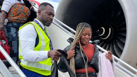 A man in a fluorescent jacket helps a woman departing a plane with her luggage
