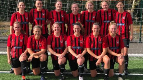 A women's football team pose for a photo in two rows. Some kneel on the front row whilst others stand behind them. All wear red and black strips.