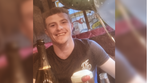 Jack Booth wearing a black t-shirt and sitting in a restaurant booth, surrounded by helium balloons. In the background there is a stained glass window, and on the table in front of him there is a full pint of beer and an opened bottle of prosecco. Jack  is clean shaven, has light brown hair which is cut shorter on the sides, and is smiling at the camera.