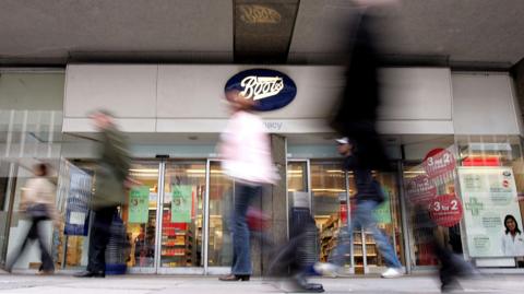 Front doors of Boots with about four blurry pedestrians walking past in the foreground.