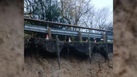 Close up shot of the side of the road with a grey fence in the foreground and trees in the background. There are orange cones by the fence too. Underneath the fence is exposed soil which shows signs of a large landslip.