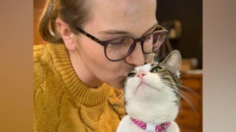 A woman wearing brown glasses and a yellow knitted jumper kisses the head of a brown and white cat that has its head slightly raised toward her. It is wearing a pink collar with white polka dots on it. 