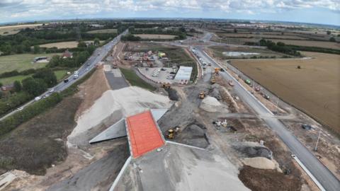 An aerial photo of a road construction site. It shows several roads, piles of sand and vehicles. There are some cars on the completed roads, and fields surround the site.