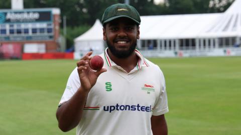 Rehan Ahmed holds a cricket ball up to the camera while smiling