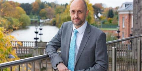 A man with very short brown hair is wearing a white shirt, blue tie and grey suit jacket. He is leaning on a balcony with a river in the background