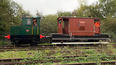Three people wearing orange hi-vis jackets stand inside an old heritage train as it travels down a track in the countryside.
