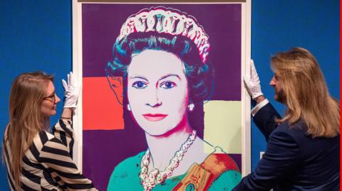 A colourful screenprint of the late Queen. She is wearing a green dress with orange sash. Two curators are holding either side of the picture. One is a man with long brown hair and a brown beard. The other is a woman who is wearing a black and white stripped top. She has long brown hair and is wearing glasses. They both have white gloves on.