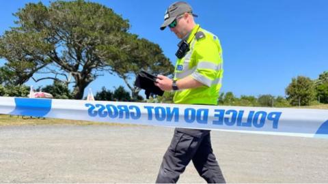 A police officer walking behind police tape with trees and blue sky in the background.