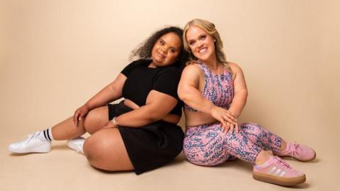 Two women sat back to back on the floor and smiling. The woman on the left is wearing black sportswear while the woman on the right is wearing pink sportswear. Both are sat against a peach background