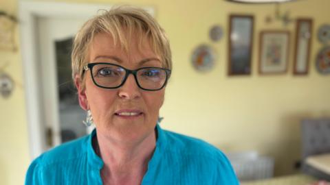 A woman stands in her kitchen. She has blonde hair, glasses and is wearing a blue shirt. 