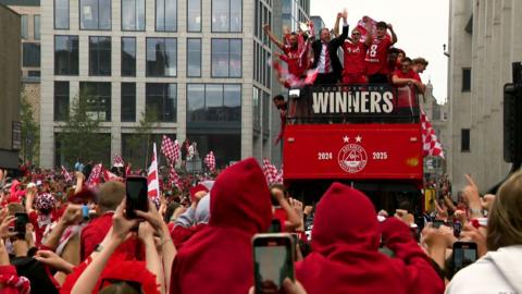 Aberdeen players and manager stand on top of a red open-topped bus holding a silver trophy while supporters celebrate.