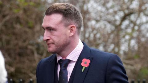 Stuart Hogg wearing a suit and tie wearing a poppy looks off to the left with trees in the background