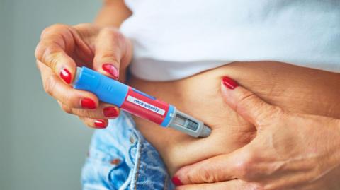 A woman, wearing bright red nail polish and unbuttoned blue jeans, injects herself into the skin and soft tissue of her lower abdomen with an obesity jab pen.