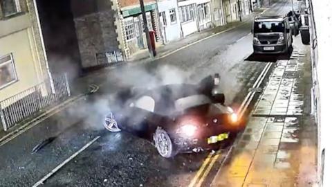 A smoking vehicle has come to a stop in middle of a road after crashing into a wall. There is smoke rising from both the inside and outside of the car and there is debris on the floor 