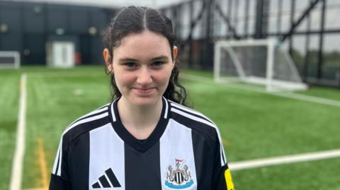 Holly, who is 15, is standing on artificial football turf with a goal, out of focus, in the background. She's wearing a NUFC home shirt and has fair skin and long dark curly hair that's tied in a ponytail. She is smiling. 