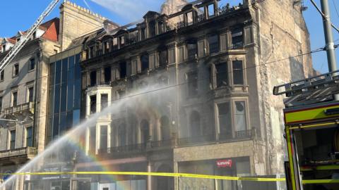 A large historic multi-storey building with extensive fire damage, particularly on the upper floors and roof. A fire hose is spraying water on the building.