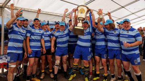 A team photograph of Hospice Barbarians, who wear a striped blue kit with the logo on the top. The team are celebrating together and holding a cup in a marquee.