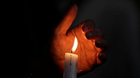 A hand shielding a candle flame above a white candle against a dark background