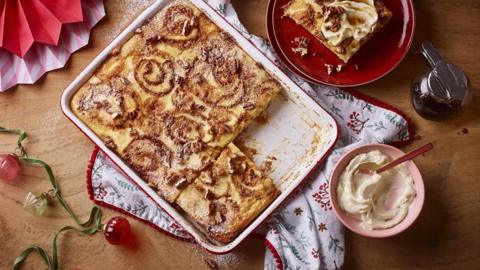 A cinnamon roll pancake traybake sits on a table alongside whipped cream
