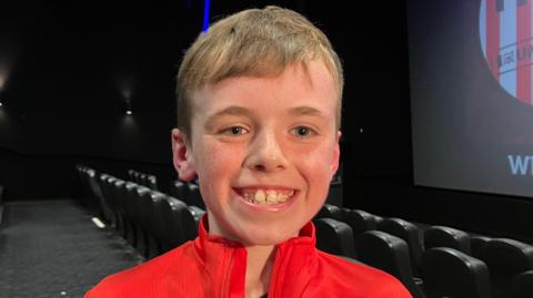 Logan is seen smiling from ear to ear inside a cinema. 
He has short hair and is wearing a Derry City jacket. 
A number of empty seats are seen behind him. 