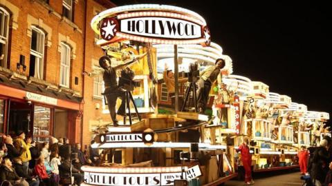 Revellers on floats entertain the crowds as they take part in the Procession through the town during the Bridgwater Guy Fawkes Carnival. There is a large, fully illuminated float coming down the street, which says Hollywood at the front top and Studio Tours at the front bottom of it. There are performers hanging off it and crowds outside shops cheering and clapping their hands.