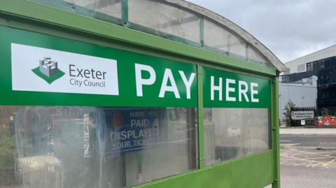 A sign saying 'pay here' next to the Exeter City Council sign on a glass and metal structure housing a parking ticket machine