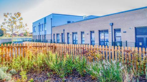 A school building with a wooden picket fence and plants and a tree in front. 